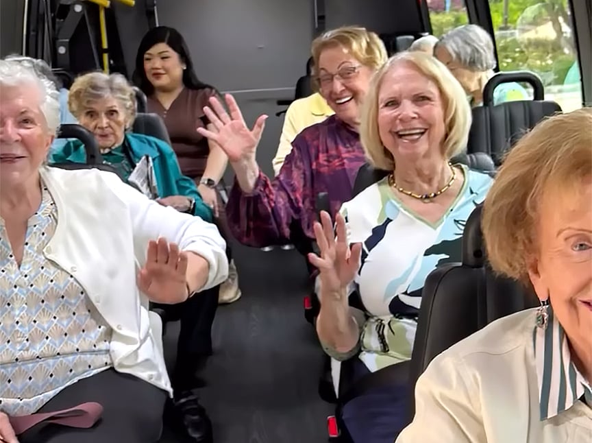 A group of older women are smiling and waving while seated inside a small bus or van. They appear cheerful and are looking toward the camera. The vehicle is well-lit and several women are sitting in the back.