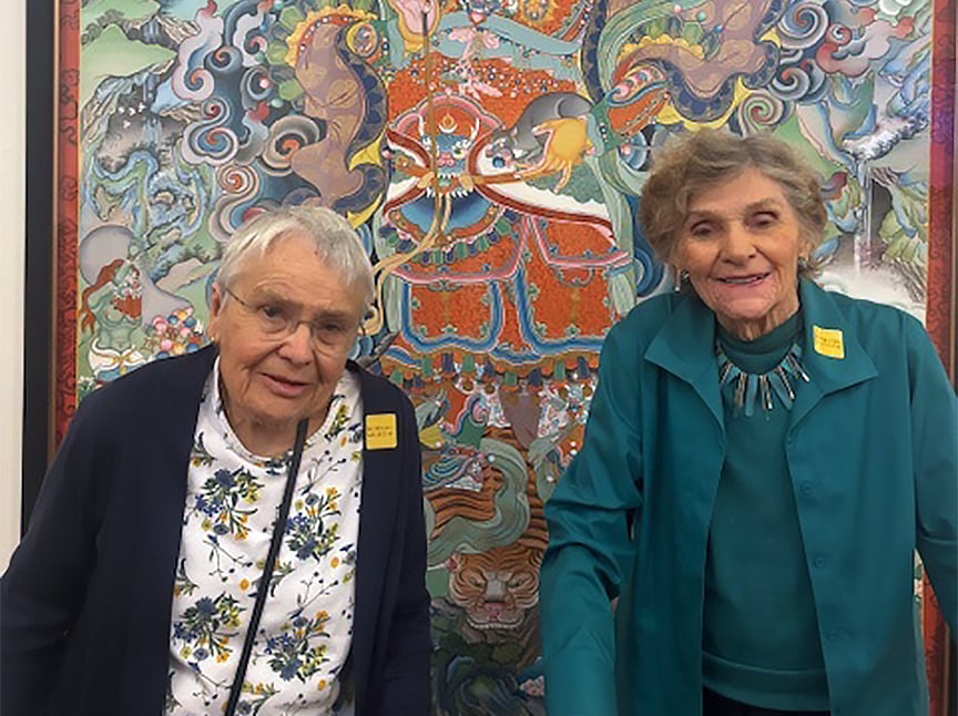 Two elderly women stand in front of a vibrant, colorful tapestry featuring intricate patterns and mythical figures. Both women wear glasses and name tags, smiling warmly at the camera.