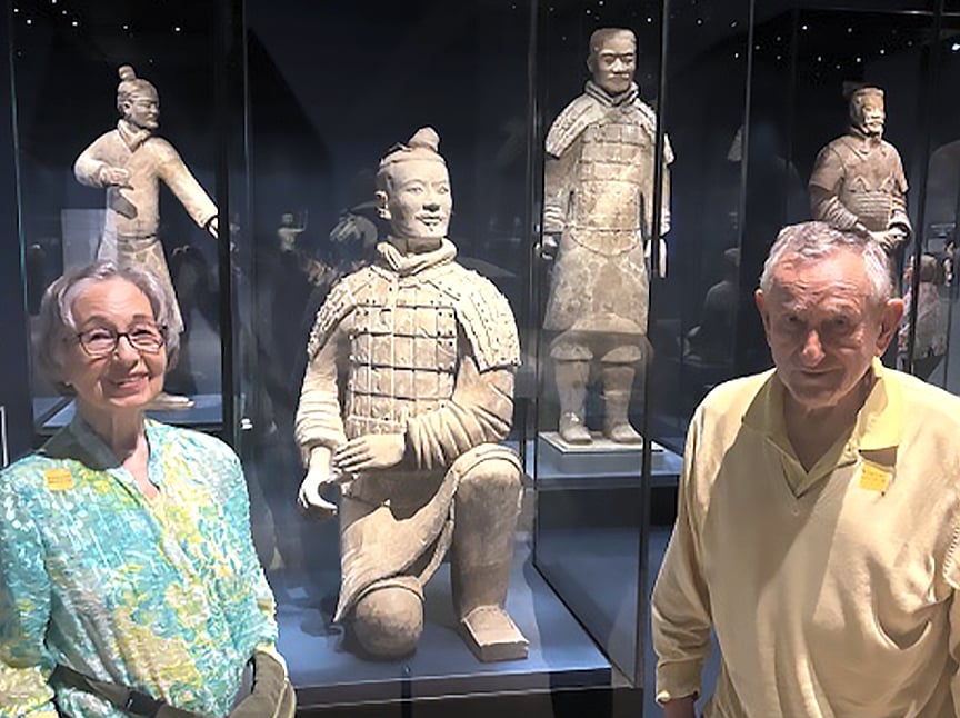 An older woman and man stand smiling in front of glass cases displaying life-sized Terracotta Warrior statues in a museum exhibit.