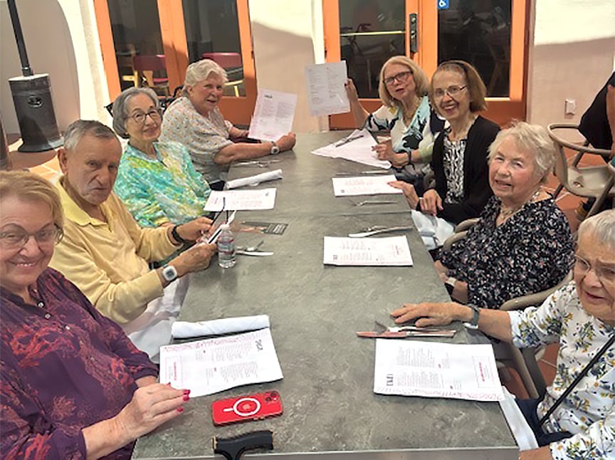 A group of elderly people, mostly women, sit around an outdoor table at a restaurant, holding menus and smiling at the camera. The weather appears sunny and pleasant.