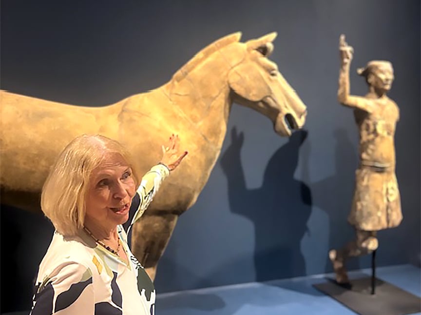 A woman gestures and smiles next to a large ancient horse statue and a human warrior statue in a museum exhibit. The background is dark blue, highlighting the sculptures.