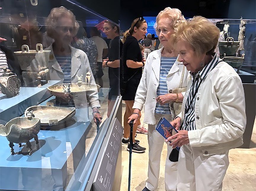 Two elderly women look at ancient artifacts displayed in a glass case at a museum, with other visitors visible in the background. One woman holds a brochure and both appear engaged with the exhibit.