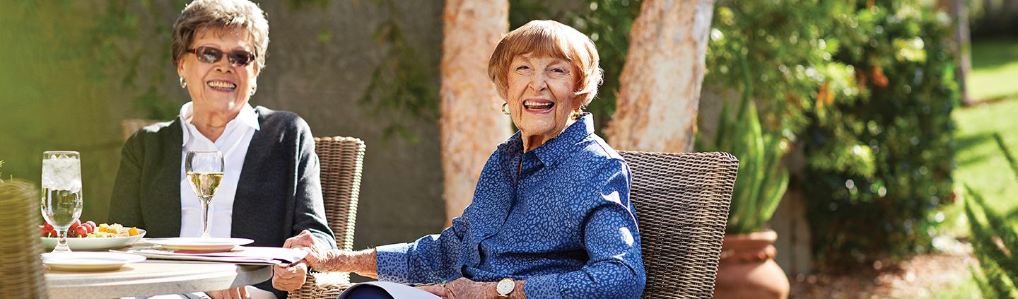 Two elderly women sit outdoors at a table, smiling and enjoying drinks and snacks in a sunny garden setting. One woman wears sunglasses, and both appear cheerful and relaxed.