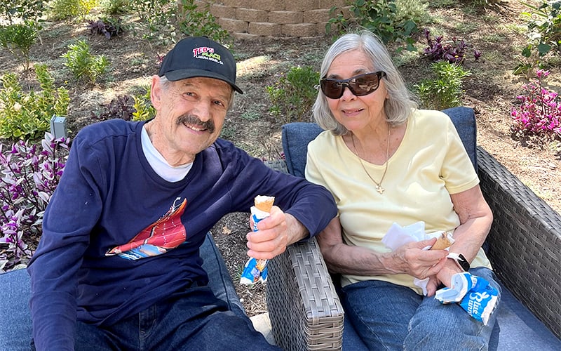 An older man and woman sit outdoors on patio chairs, smiling and enjoying ice cream bars. The man wears a dark cap and shirt, while the woman wears sunglasses and a light yellow top. Green plants and purple flowers are in the background.