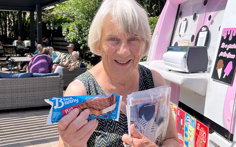 An older woman smiles while holding a Blue Bunny ice cream sandwich and a magazine, standing outside near an ice cream truck with people sitting in the background.