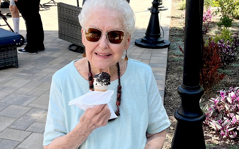 Elderly woman with short white hair and sunglasses smiles while holding an ice cream cone outdoors. She wears a light blue shirt and a beaded necklace, sitting on a patio surrounded by plants and lamp posts.