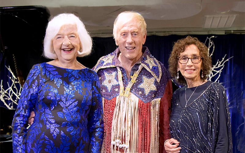 Three older adults stand together, smiling. The woman on the left wears a blue dress with a leafy pattern. The man in the center wears a star-patterned, fringed jacket. The woman on the right wears a navy dress with sparkly details.