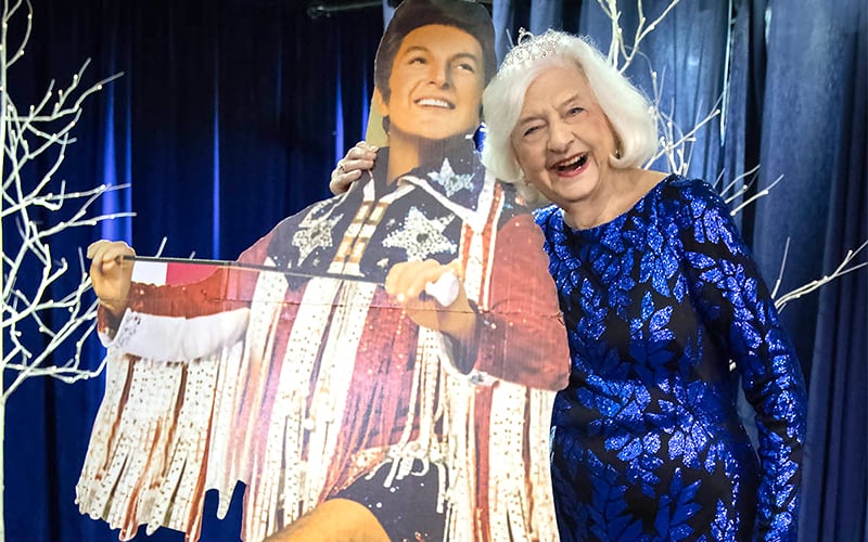 An elderly woman in a blue floral dress and tiara smiles while posing with a life-sized cardboard cutout of an exuberant performer in a glittery costume. White decorative branches and blue curtains are in the background.