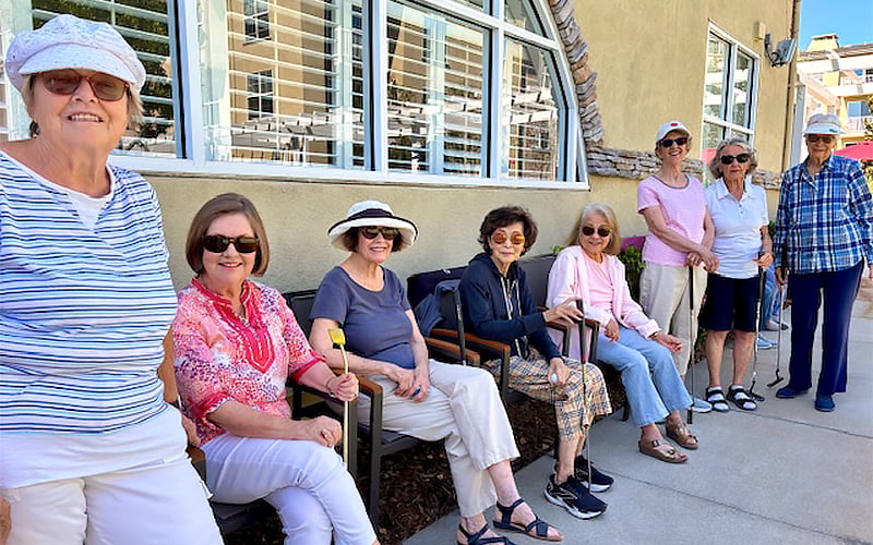 Eight older women in casual clothes and sun hats sit and stand outside a building on a sunny day, smiling at the camera. Some are seated on a bench, while others stand on the sidewalk.