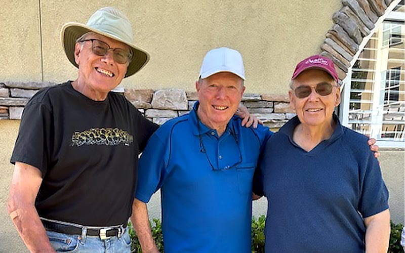 Three older men stand close together smiling outdoors. One wears a wide-brimmed hat and black shirt, another wears a white cap and blue shirt, and the third wears a red cap and navy shirt. They stand in front of a light-colored wall.