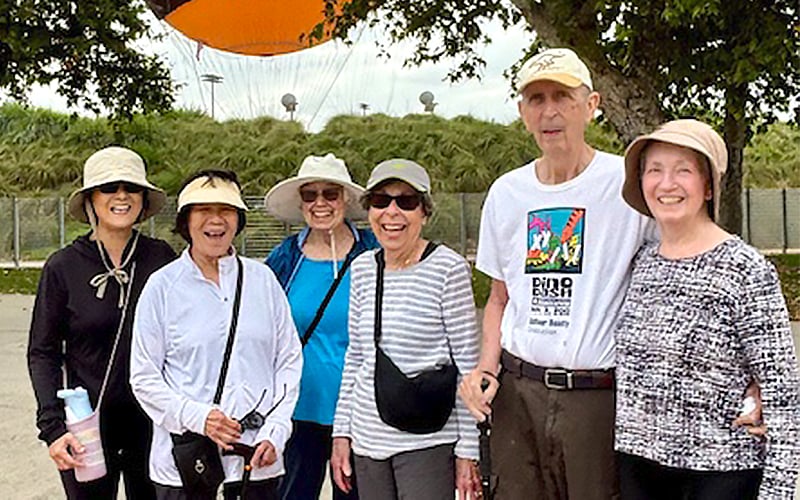 Six older adults, wearing hats and sunglasses, stand outdoors smiling together. They are dressed casually and appear to be enjoying a sunny day. Greenery and an orange balloon or structure are in the background.