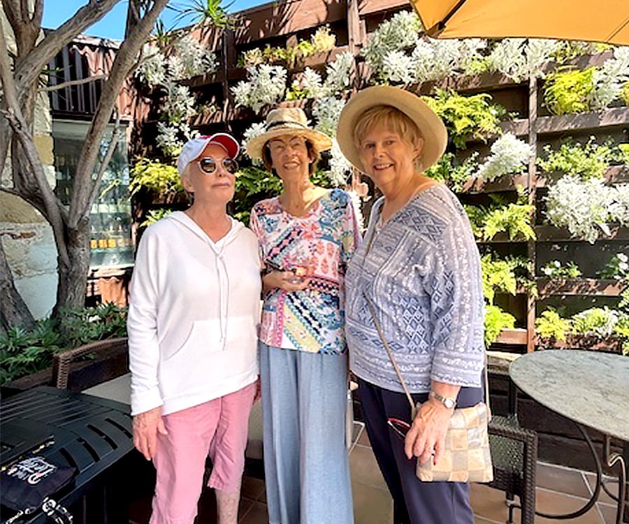 Three older women stand smiling together outdoors in front of a vertical garden wall. They wear casual clothes and hats, and sunlight filters through a nearby umbrella. Patio furniture is visible around them.