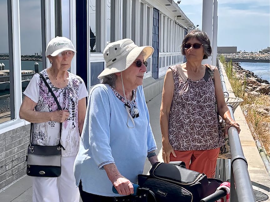 Three older women are outdoors by a waterfront on a sunny day. Two wear hats and sunglasses; one uses a walker. They are standing on a walkway next to a building with large windows.