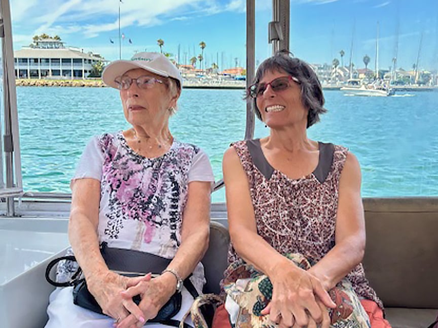 Two older women sit side by side on a boat, smiling and enjoying a sunny day on the water. Sailboats and palm trees are visible in the background, suggesting they are near a marina.