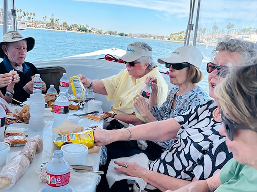 A group of older adults sit around a table on a boat, enjoying snacks and bottled water, with a scenic waterfront and palm trees visible in the background on a sunny day.
