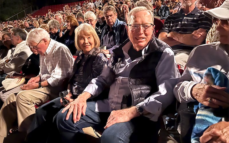 A large audience sits closely together in a theater. In the foreground, an older couple smiles at the camera while people around them chat or look toward the stage. The atmosphere appears lively and sociable.
