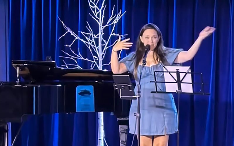 A woman in a blue dress stands at a microphone on stage, gesturing with her hands. There is a piano to her left and a white decorative tree behind her, with blue curtains as the backdrop.