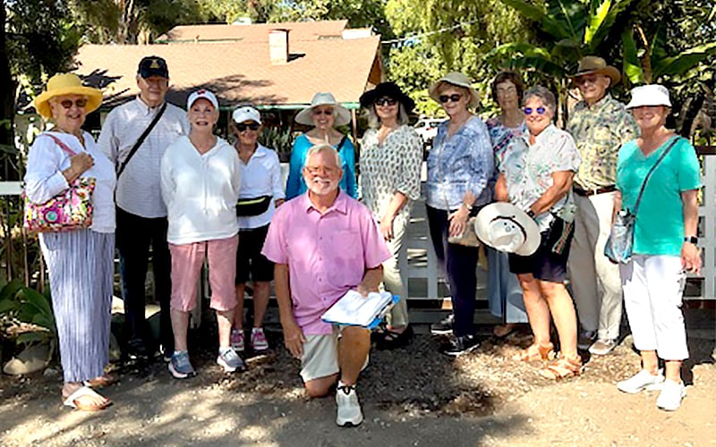 A group of thirteen older adults, some wearing hats and sunglasses, pose and smile outdoors in front of a house surrounded by trees and plants. One man kneels in front holding papers, while the rest stand closely together.
