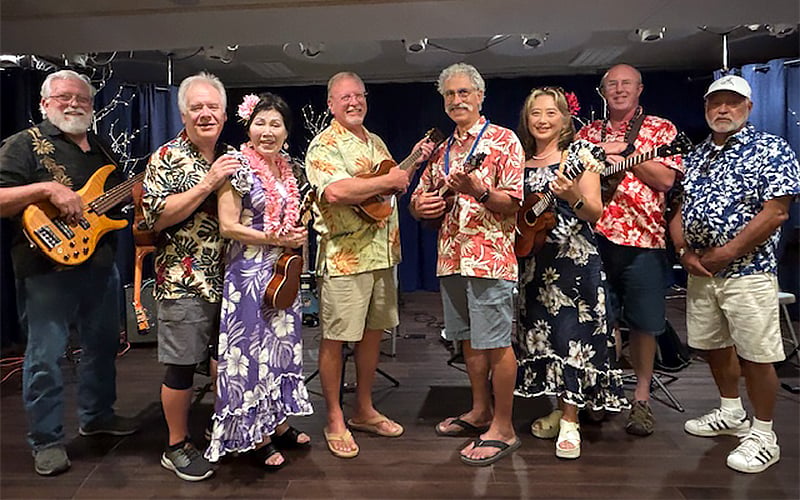 A group of eight people stand on a stage, smiling and holding musical instruments including ukuleles and a bass guitar. They are dressed in colorful Hawaiian shirts and leis, with a dark curtain as the background.