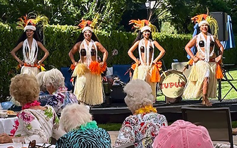 Four hula dancers in grass skirts and feathered headpieces perform on an outdoor stage, while elderly audience members watch from tables decorated with leis and flowers. A drum with TUPUA is visible in the background.