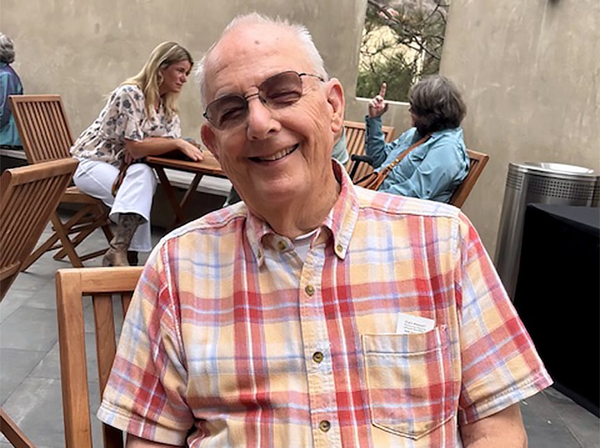 An elderly man wearing glasses and a plaid short-sleeve shirt smiles while sitting on a wooden chair outdoors, with a few people conversing at tables in the background.