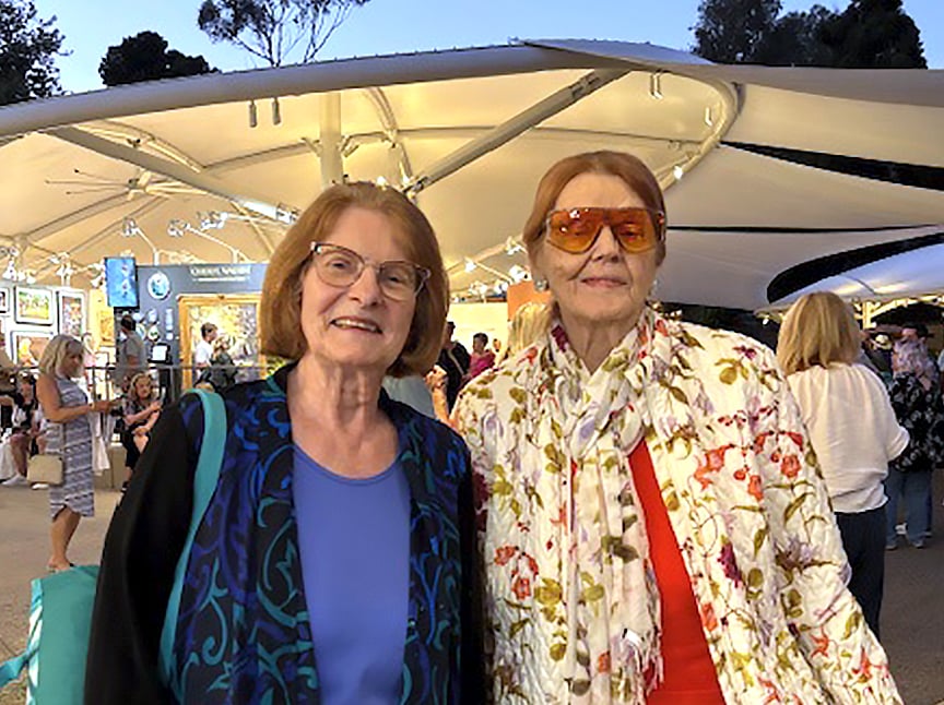 Two older women stand outdoors at an art fair. One wears glasses, a blue shirt, and a patterned vest; the other wears orange sunglasses, a floral scarf, and a red top. People and art displays are visible in the background.