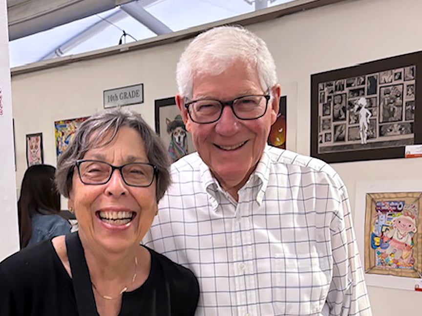 An older woman and man, both wearing glasses and smiling, stand together in front of a wall displaying various pieces of artwork.