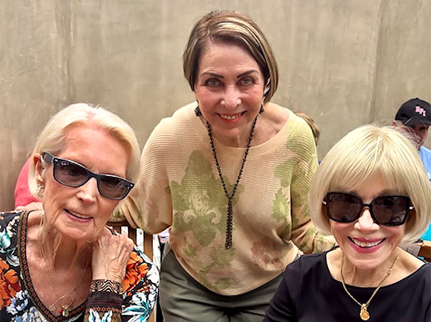 Three older women pose together and smile; two are seated wearing sunglasses, and one stands behind them. All appear happy and are dressed in casual, stylish clothing against a neutral wall background.