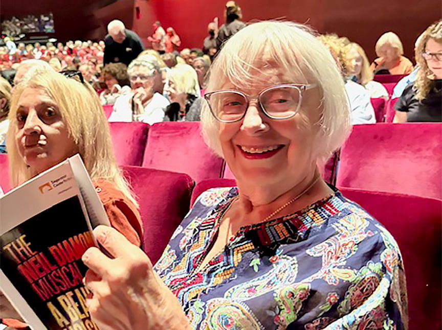 An older woman with short white hair and glasses smiles while holding a theater program in a crowded auditorium. Other audience members sit nearby on red velvet seats, waiting for a performance to begin.