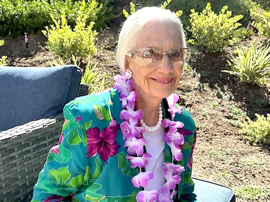 An elderly woman with white hair and glasses sits outdoors on a blue chair, smiling. She wears a teal floral jacket, white beaded necklace, and a purple flower lei. Green plants and sunlight are in the background.