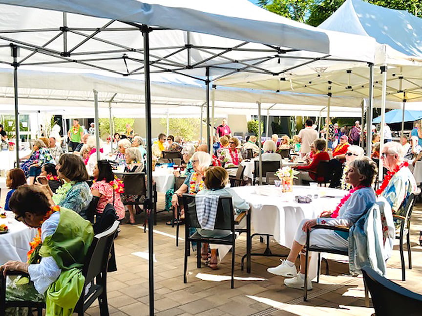 A large group of seniors sit at round tables under white canopies at an outdoor event. Many people wear colorful leis and summery clothing, with trees and sunshine in the background.