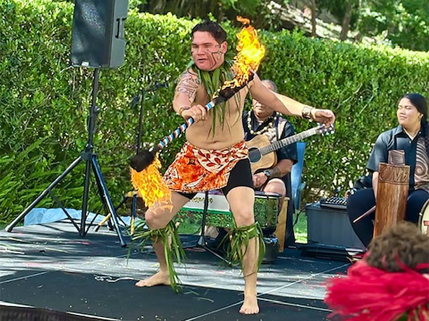 A performer wearing traditional attire spins a flaming staff on an outdoor stage, with musicians playing instruments and greenery in the background.