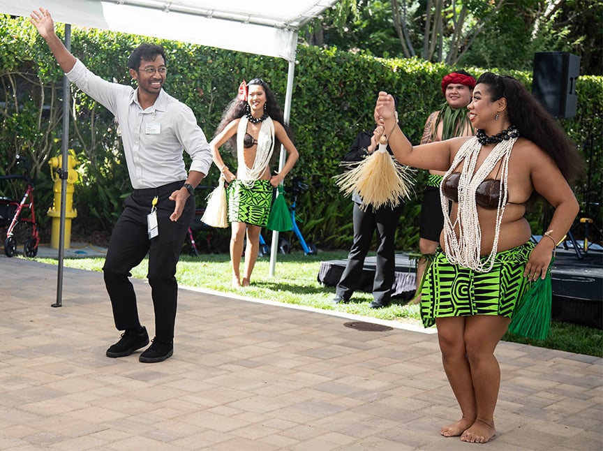 A man in business attire dances with a smiling woman in traditional Polynesian dress, with two other dancers in similar attire performing behind them outdoors under a canopy.