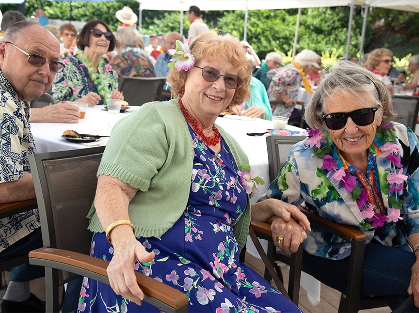 A group of smiling seniors sit at outdoor tables, dressed in colorful, tropical-themed clothing and leis, enjoying a festive event under a canopy. Two women wear flower accessories and sunglasses in the foreground.