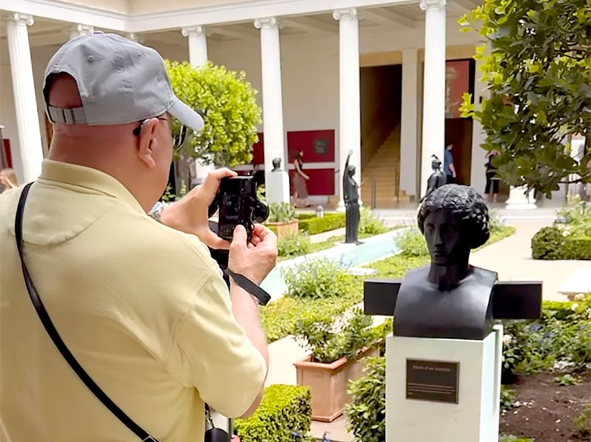 A man in a light yellow shirt and gray cap takes a photo of a black marble bust in an outdoor museum courtyard with columns, statues, and greenery.