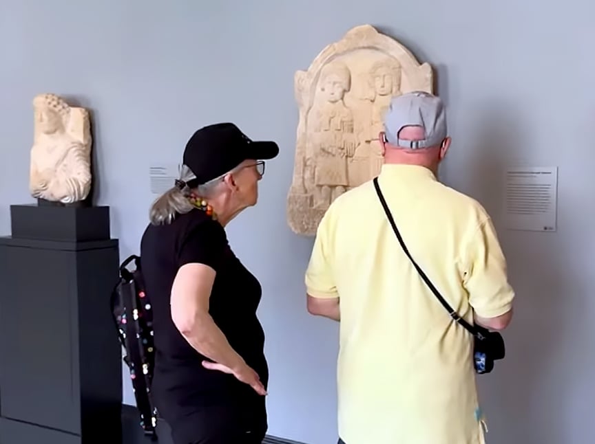 Two older adults, one in a yellow shirt and the other in black, stand closely together looking at an ancient stone relief displayed on a museum wall. Another carved artifact is visible on a pedestal nearby.