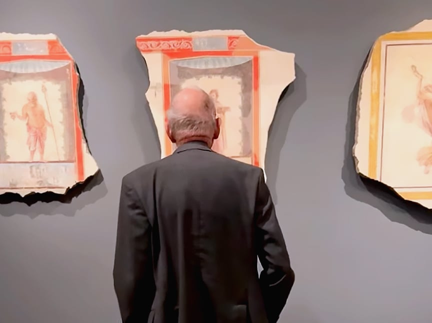 An older man in a suit stands with his back to the camera, closely viewing ancient fresco fragments displayed on a gray museum wall.