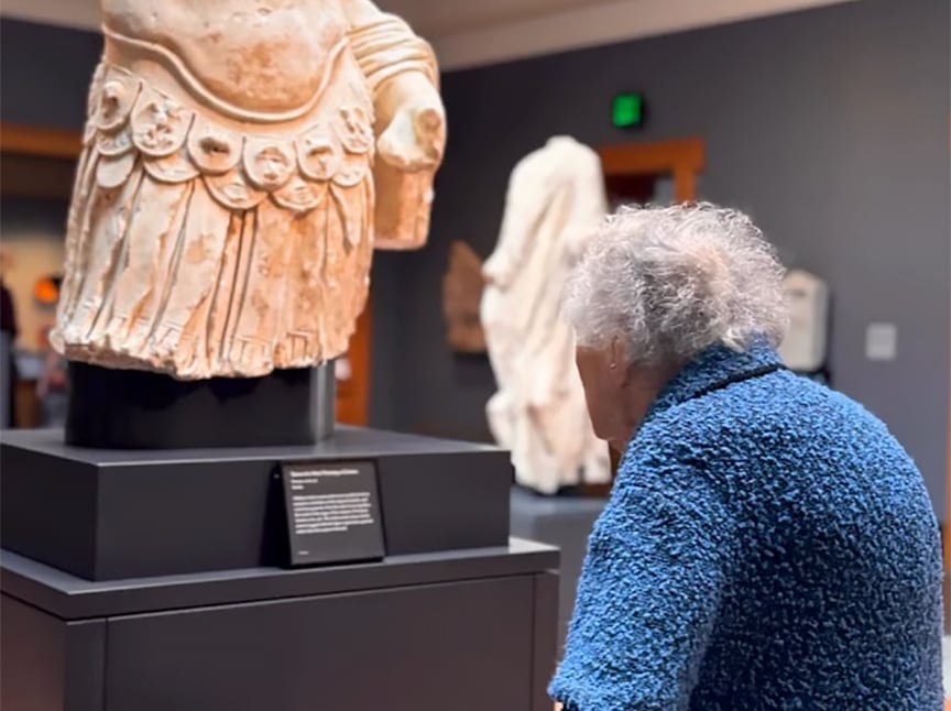 An elderly person in a blue textured jacket views an ancient marble statue displayed on a pedestal in a museum gallery. The statue is missing its head and arms.