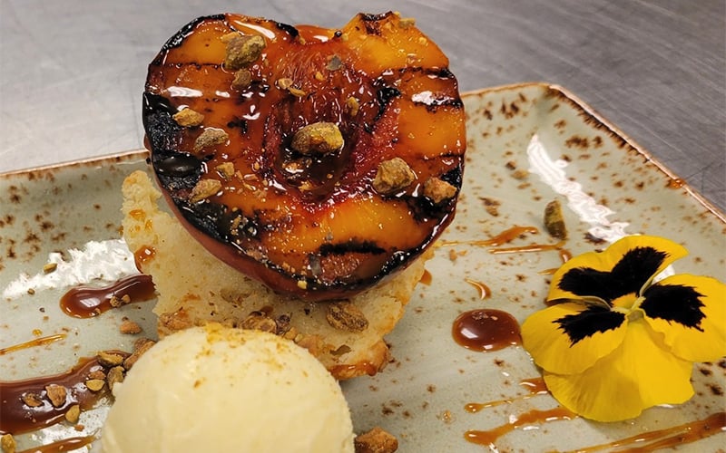 A plated dessert featuring a grilled peach half on cake, garnished with crushed nuts, caramel drizzle, a scoop of vanilla ice cream, and a yellow edible flower on a speckled ceramic plate.
