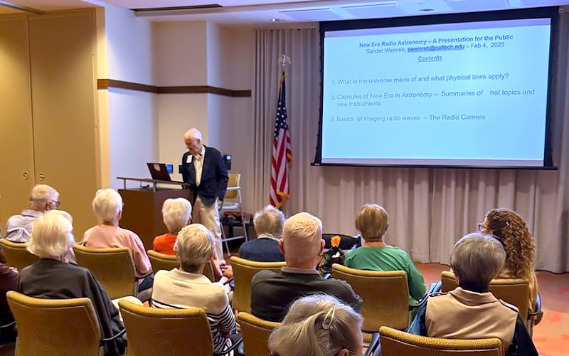 A man gives a presentation to a seated group of older adults in a conference room. A projection screen displays a talk titled New Era Radio Astronomy – A Presentation for the Public. An American flag is in the background.