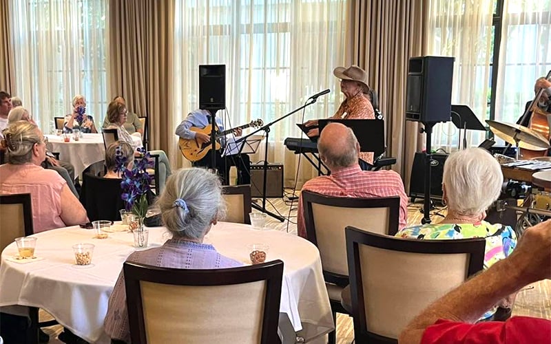 A group of elderly people sits at round tables, watching a woman in a cowboy hat sing and play keyboard, accompanied by a small band with guitar and drums in a sunlit room with large windows and beige curtains.