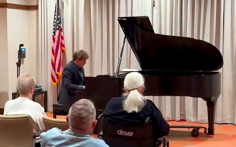 A pianist performs on a grand piano in front of an audience. Three people, including one in a wheelchair, watch. An American flag stands in the background, and light-colored curtains cover the wall behind the piano.