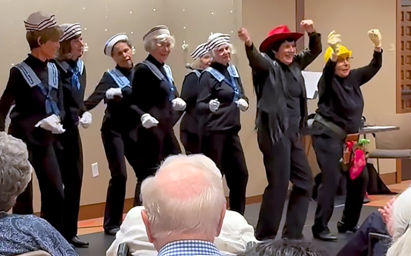 A group of older women perform on stage, most dressed in matching sailor outfits, while two on the right wear black with colorful hats and props. Audience members are visible in the foreground, watching the lively performance.