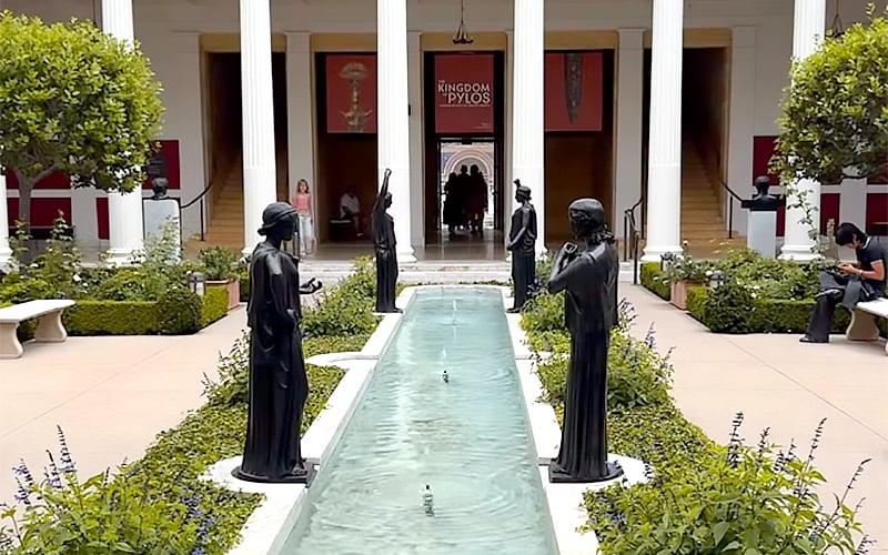 A courtyard with a central rectangular fountain, surrounded by four black statues, green plants, benches, and white columns. The backdrop features a doorway marked Kingdom of Pylos.