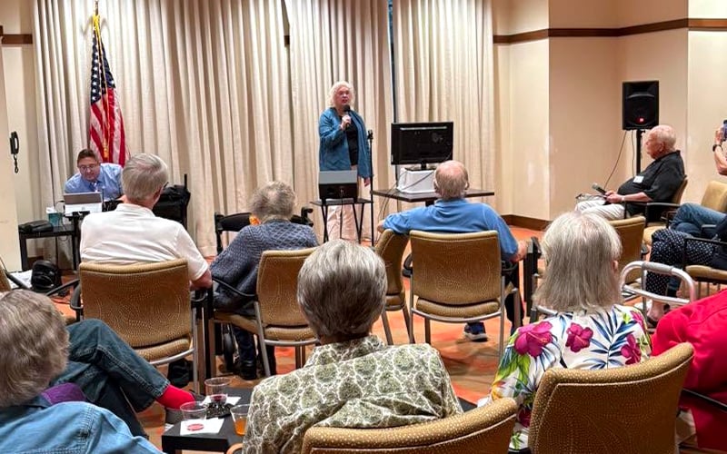 A group of people seated in chairs face a woman speaking at the front of a room with beige curtains, an American flag, and electronic equipment. The audience appears to be mostly older adults.