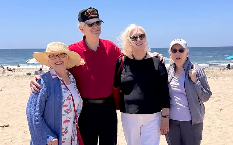 Four older adults stand together smiling on a sandy beach with the ocean and blue sky in the background. Three women wear sun hats and sunglasses, while the man wears a cap and sunglasses.