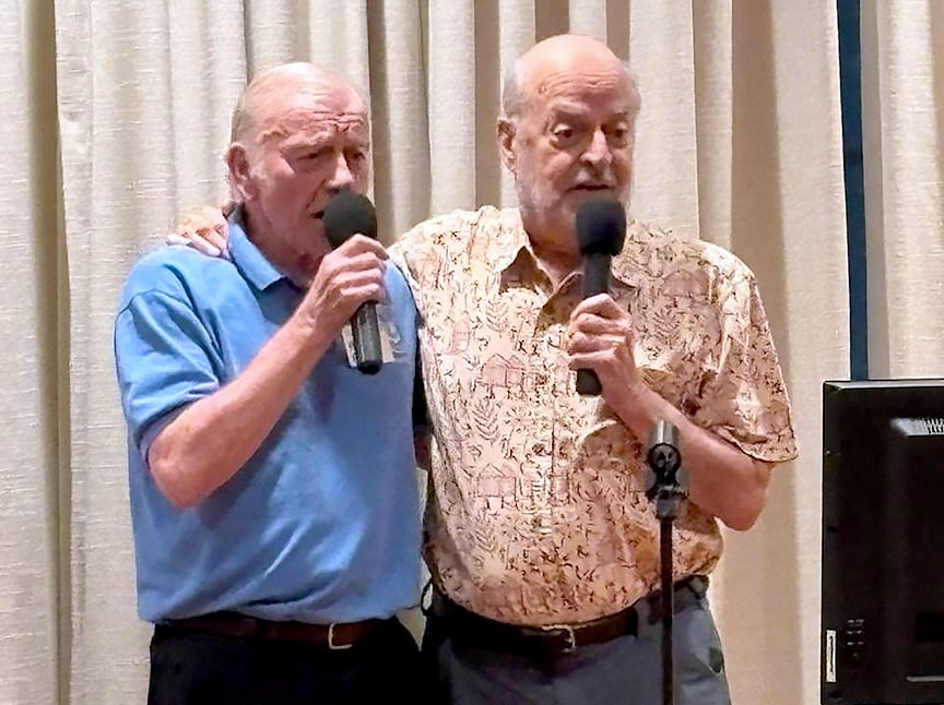 Two elderly men are standing close together, each holding a microphone and singing. One wears a blue polo shirt, the other a beige patterned shirt. Beige curtains hang in the background.