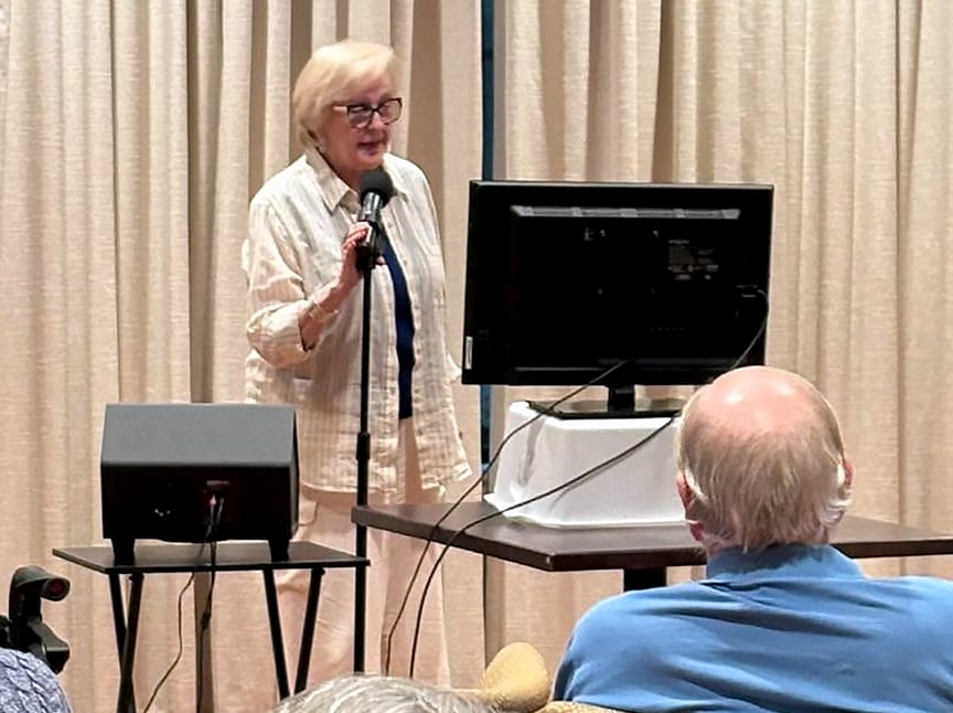 An older woman speaks into a microphone while standing beside a monitor and small speaker, addressing an audience in a room with beige curtains. A man with gray hair sits facing her in the foreground.