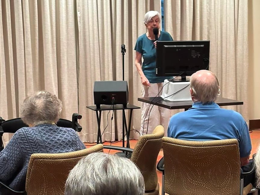 An older woman stands at a microphone in front of a monitor, speaking to a seated audience of seniors in a room with beige curtains.
