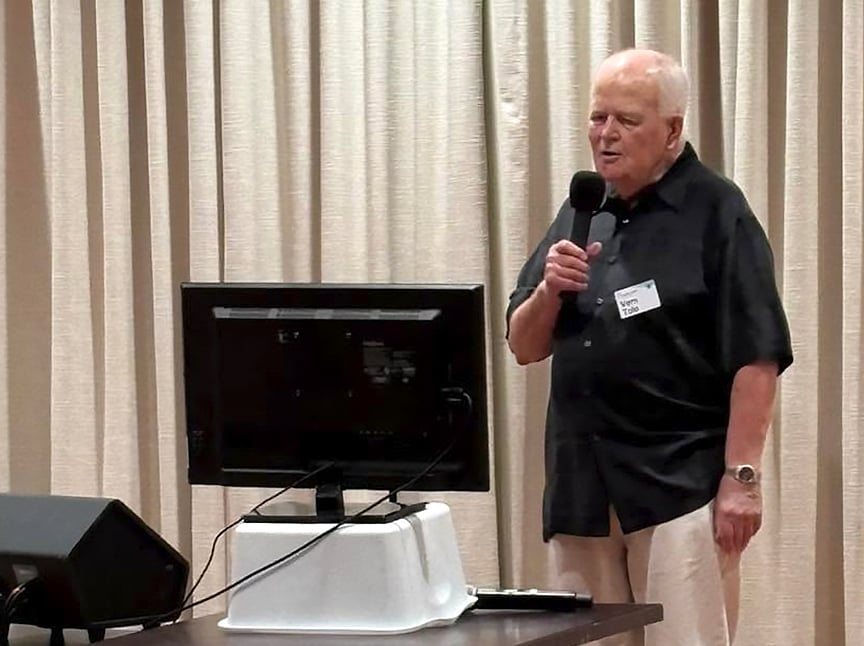 An older man in a black shirt holds a microphone and stands beside a computer monitor on a table, speaking in front of beige curtains.